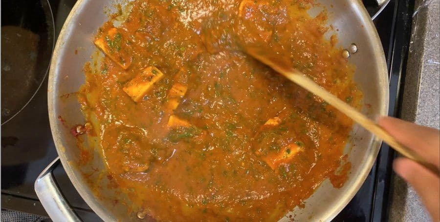 Ginger-cilantro paneer curry simmering in a white pan with a wooden spoon, showing a rich golden-brown gravy.