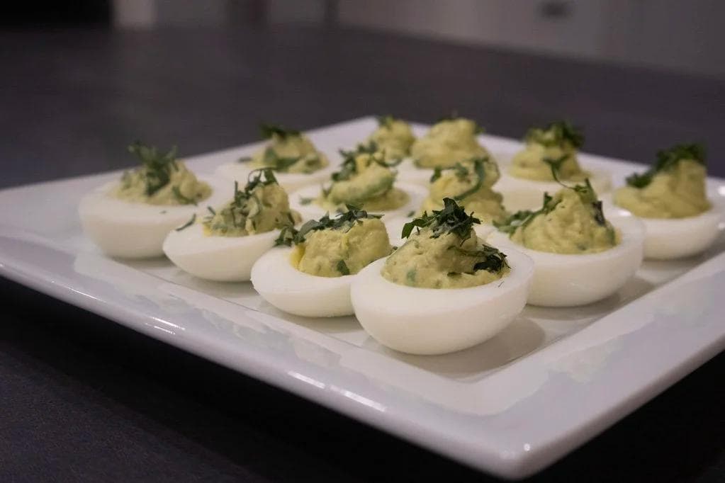 Avocado deviled eggs arranged on a white plate, topped with green avocado mixture and fresh herb garnish.