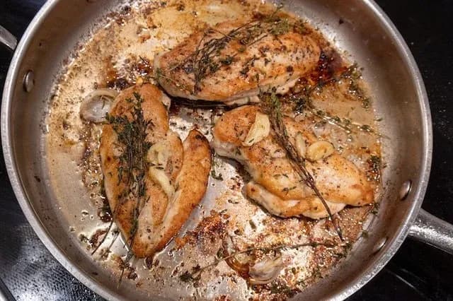 Pan-fried chicken breasts with golden-brown crust, garnished with fresh thyme sprigs in a stainless steel skillet.