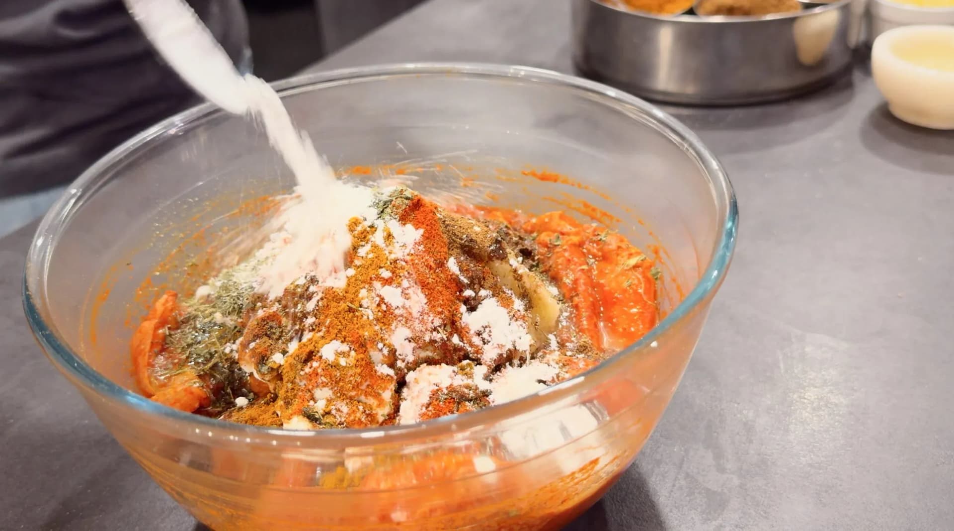 Glass bowl filled with marinated chicken being coated with spices including paprika, herbs, and salt for tikka marinade.