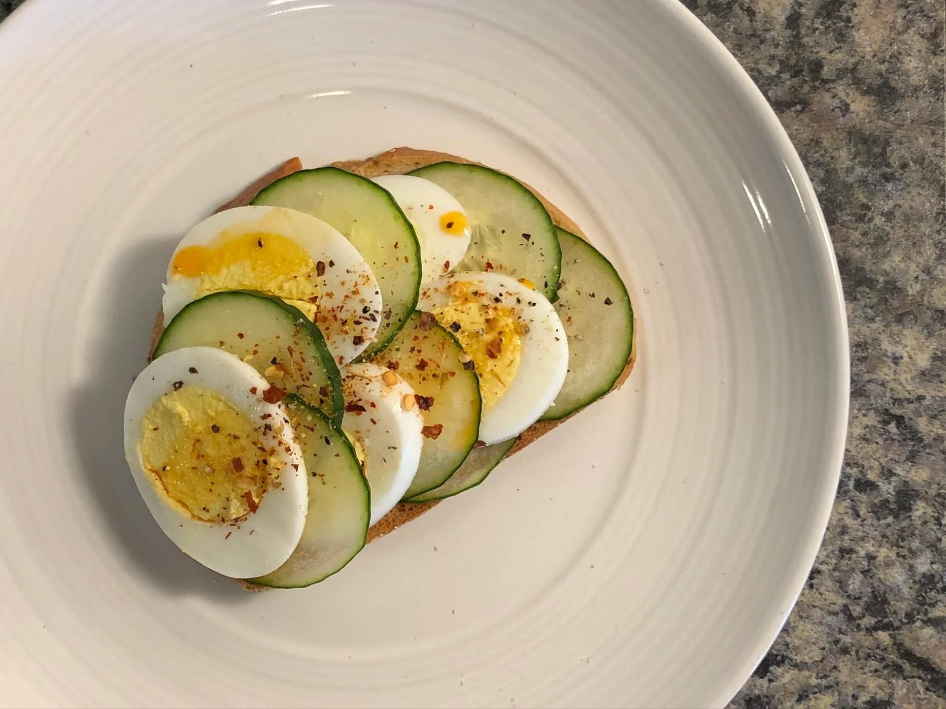Cucumber and boiled egg open-faced toast topped with sliced eggs, cucumber rounds, and red pepper seasoning on a white plate.