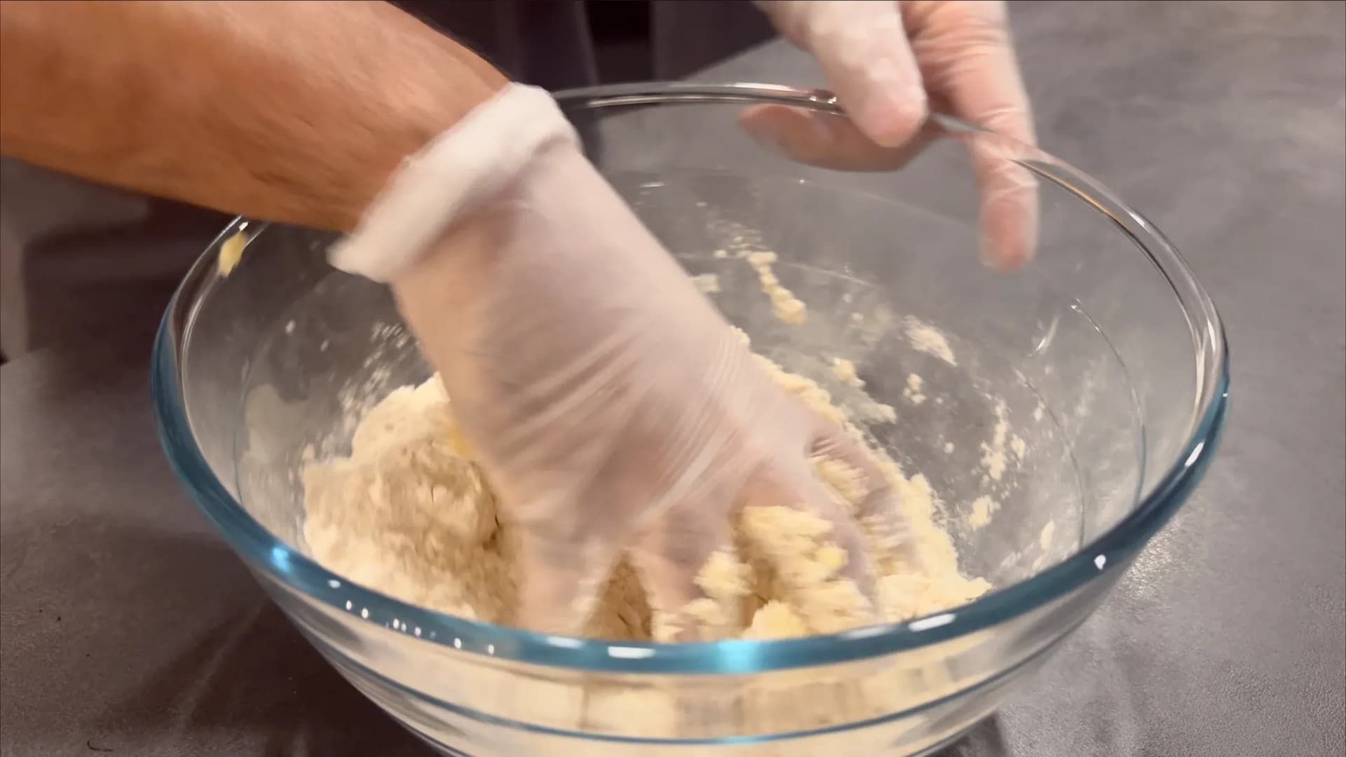 Hands in plastic gloves mixing pie dough in a glass bowl with a wooden spoon.
