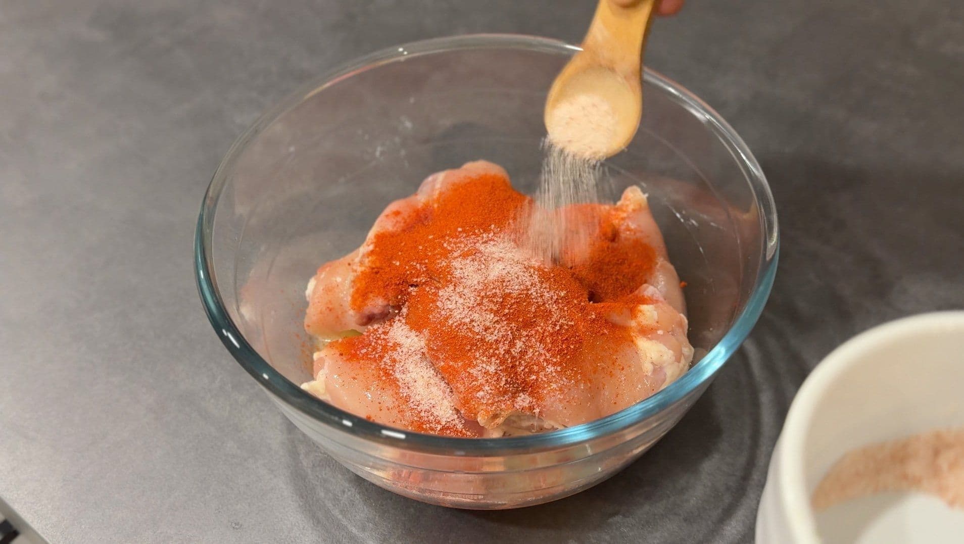 A hand brushing spices onto raw chicken pieces in a glass bowl for the first tandoori marinade.