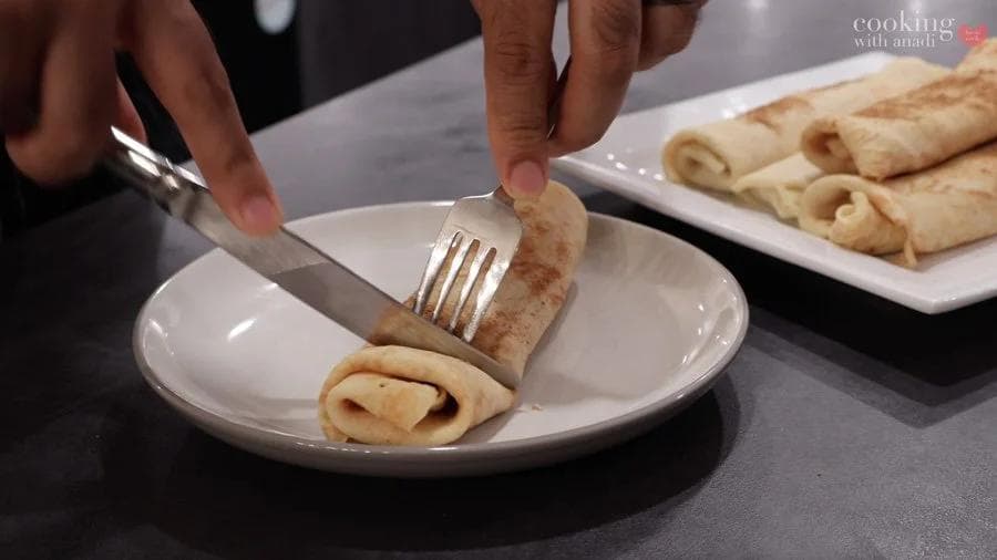 Cutting into a rolled pumpkin crepe on a white plate with a fork and knife, with more crepes on a serving plate in the background.