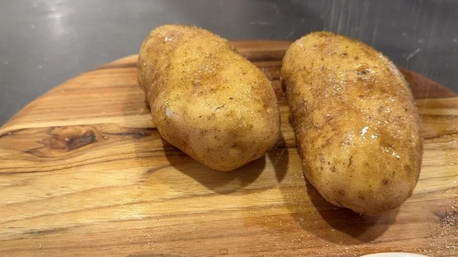 Two russet potatoes rubbed with olive oil and seasoned with salt and pepper on a wooden cutting board.