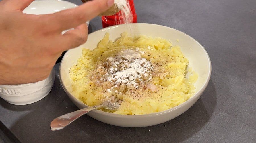 Mashed potatoes being seasoned with salt and pepper in a white bowl during llapingachos preparation.