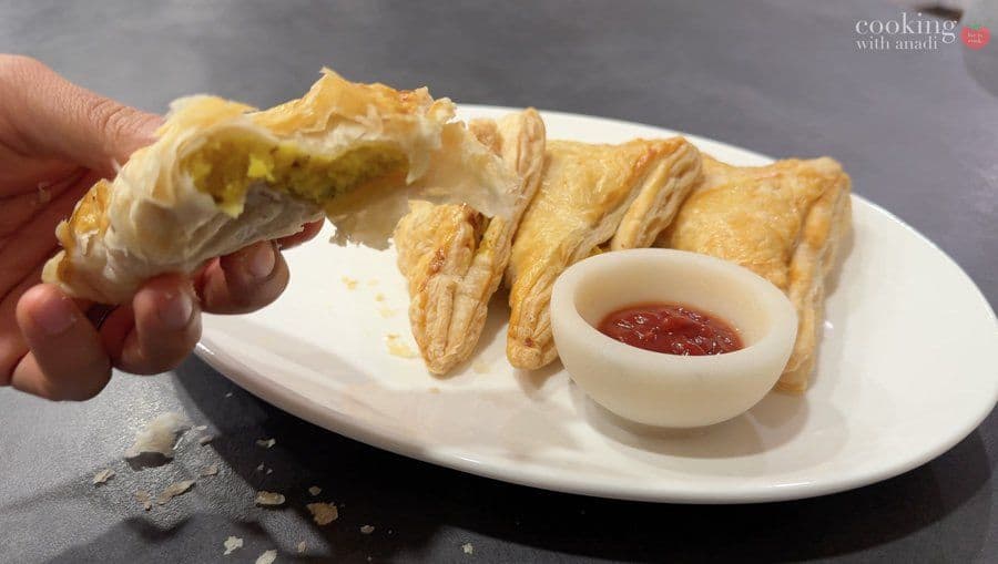 Hand holding a golden, flaky aloo patty with potato filling, served with ketchup on a white plate.