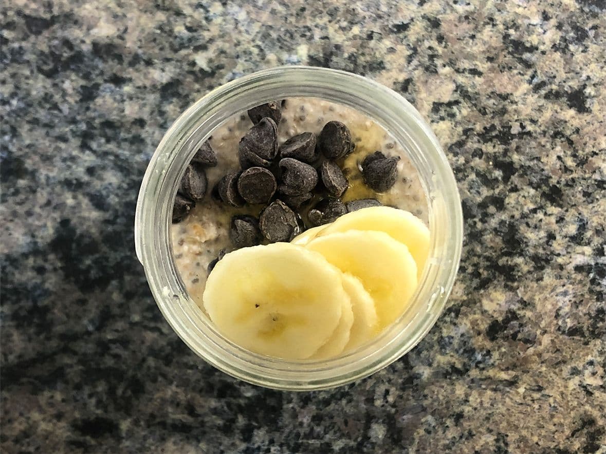 Chocolate chips and banana slices in a clear container with oat mixture for overnight oats.