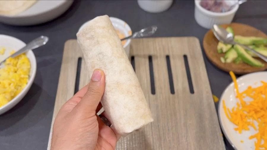 Hand holding a rolled breakfast burrito with visible filling on a wooden cutting board surrounded by toppings.