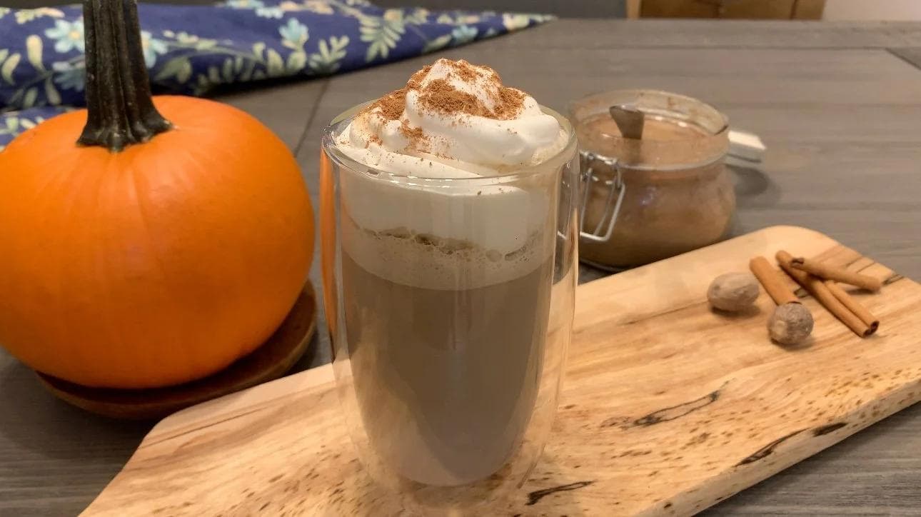 Homemade pumpkin spice latte in a glass topped with whipped cream and cinnamon, beside a pumpkin and spices on a wooden board.