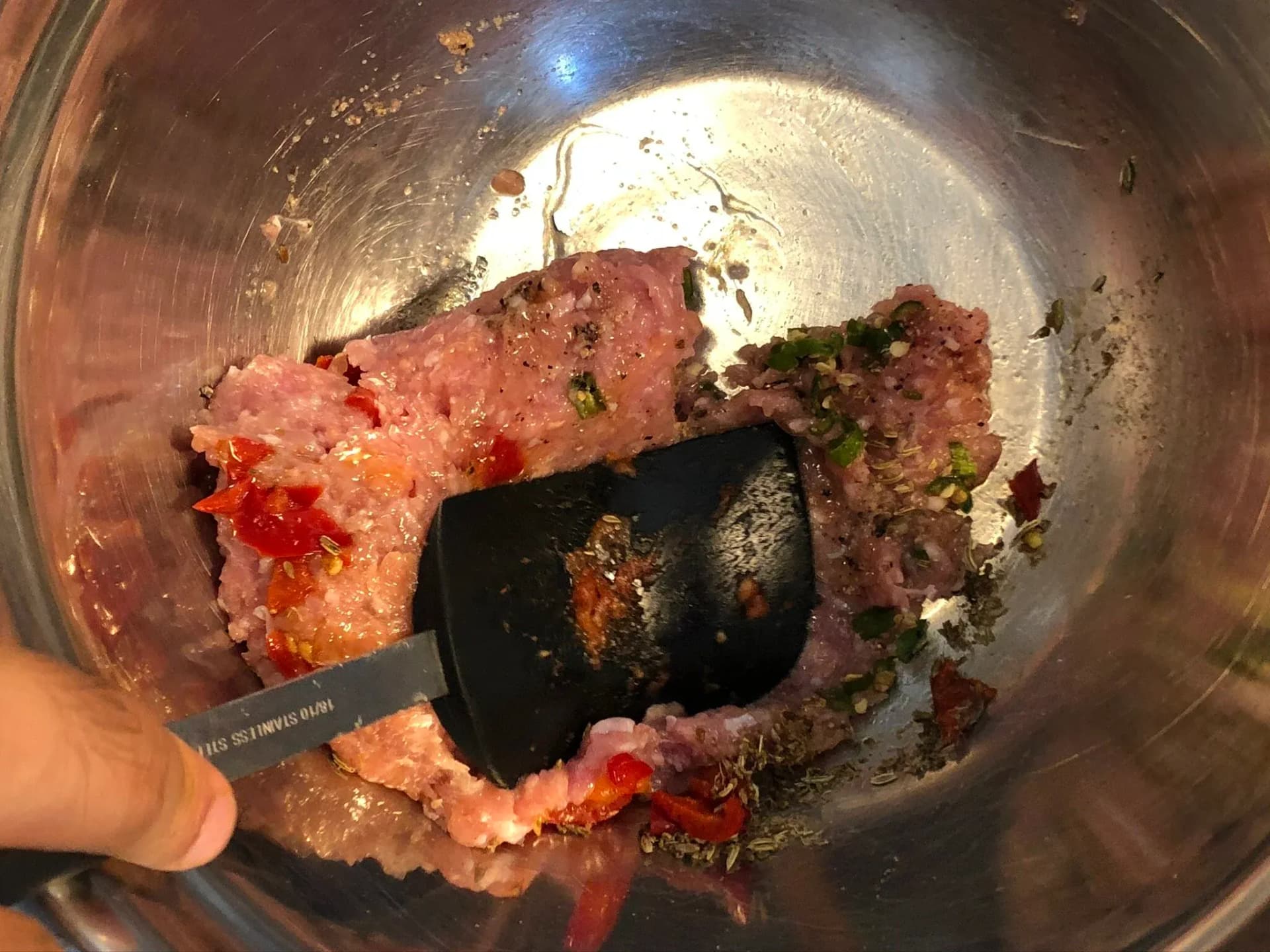 Mixing ground meat with red peppers and herbs in a metal bowl with a black spatula.