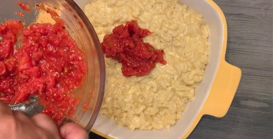 Pouring tomato sauce over cooked mac and cheese in a yellow casserole dish for Pizza Mac and Cheese.