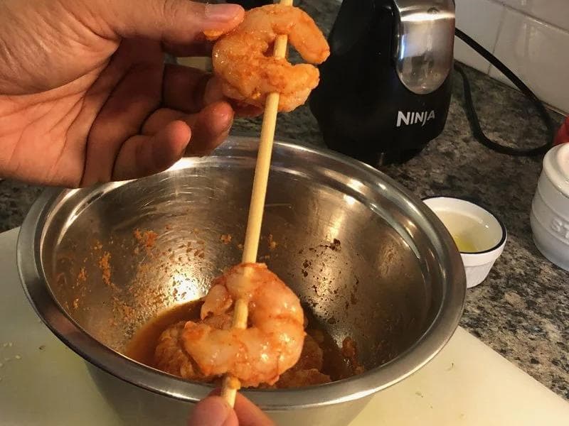 Shrimp threaded on wooden skewers being lifted from a bowl of orange-red tandoori marinade