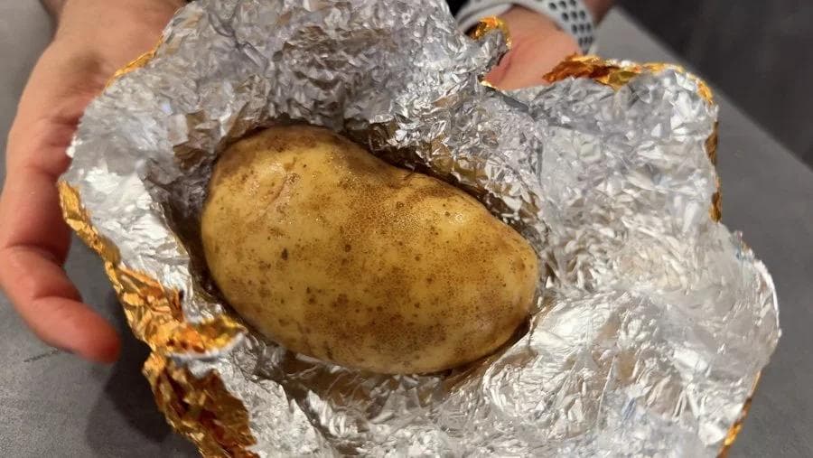 Baked potato wrapped in aluminum foil being held in hands, showing soft skin after air frying without foil.
