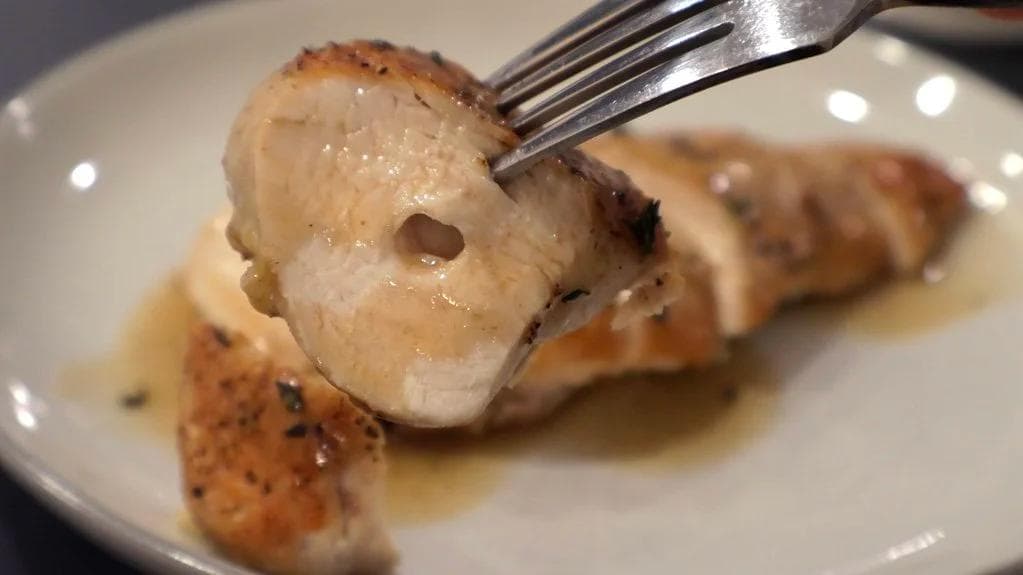 Sliced pan-fried chicken breast with golden-brown skin on a white plate with pan sauce.