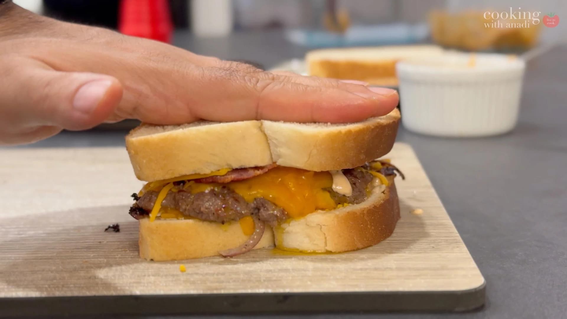 Hand pressing down on a smash burger patty melt with melted cheddar cheese and bacon between toasted bread slices.