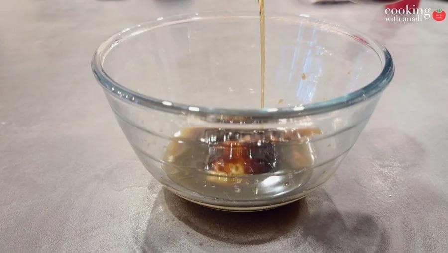 Sesame oil being poured into a glass bowl containing soy sauce, oyster sauce, and other dark marinade ingredients.