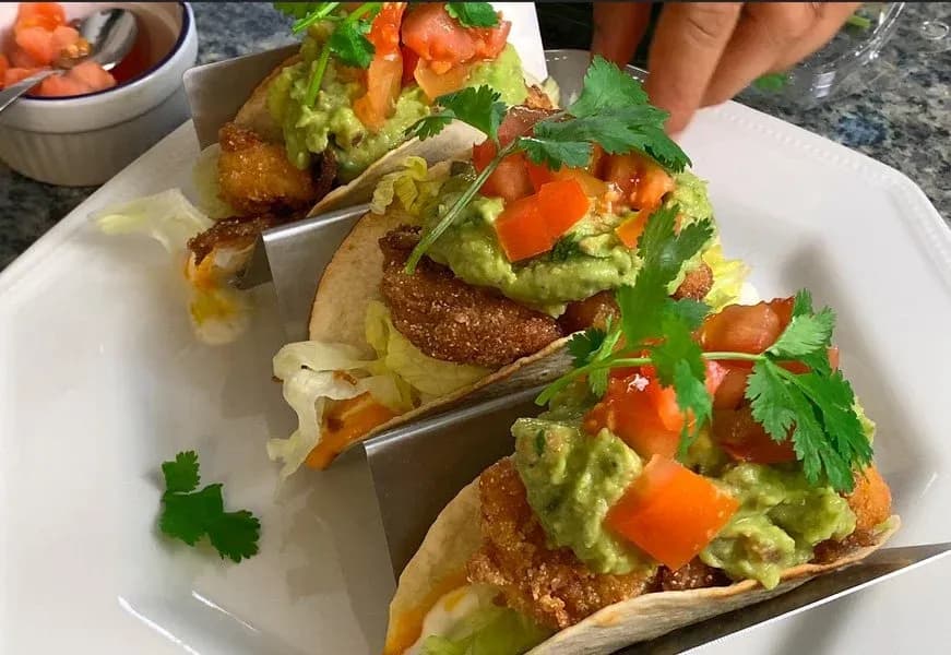 Crispy chicken and shrimp tacos topped with guacamole, diced tomatoes, and fresh cilantro on a white plate.