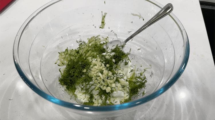 Mixed tzatziki ingredients in a glass bowl with shredded cucumber, dill, and garlic before chilling.