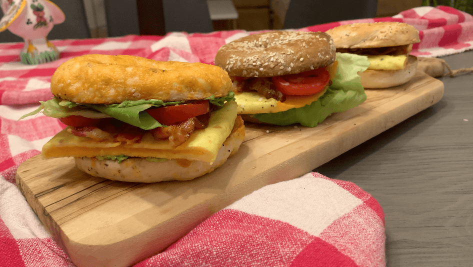 Three breakfast bagel sandwiches on a wooden board with lettuce, tomato, cheese, and bacon on a red checkered tablecloth.