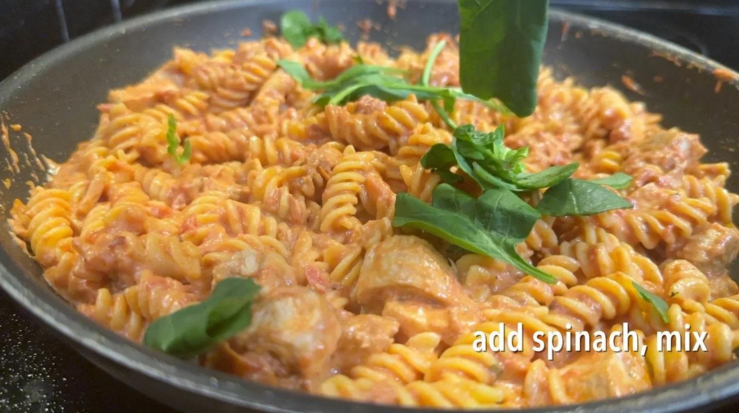 Creamy tomato pasta with diced chicken and fresh spinach being mixed in a skillet
