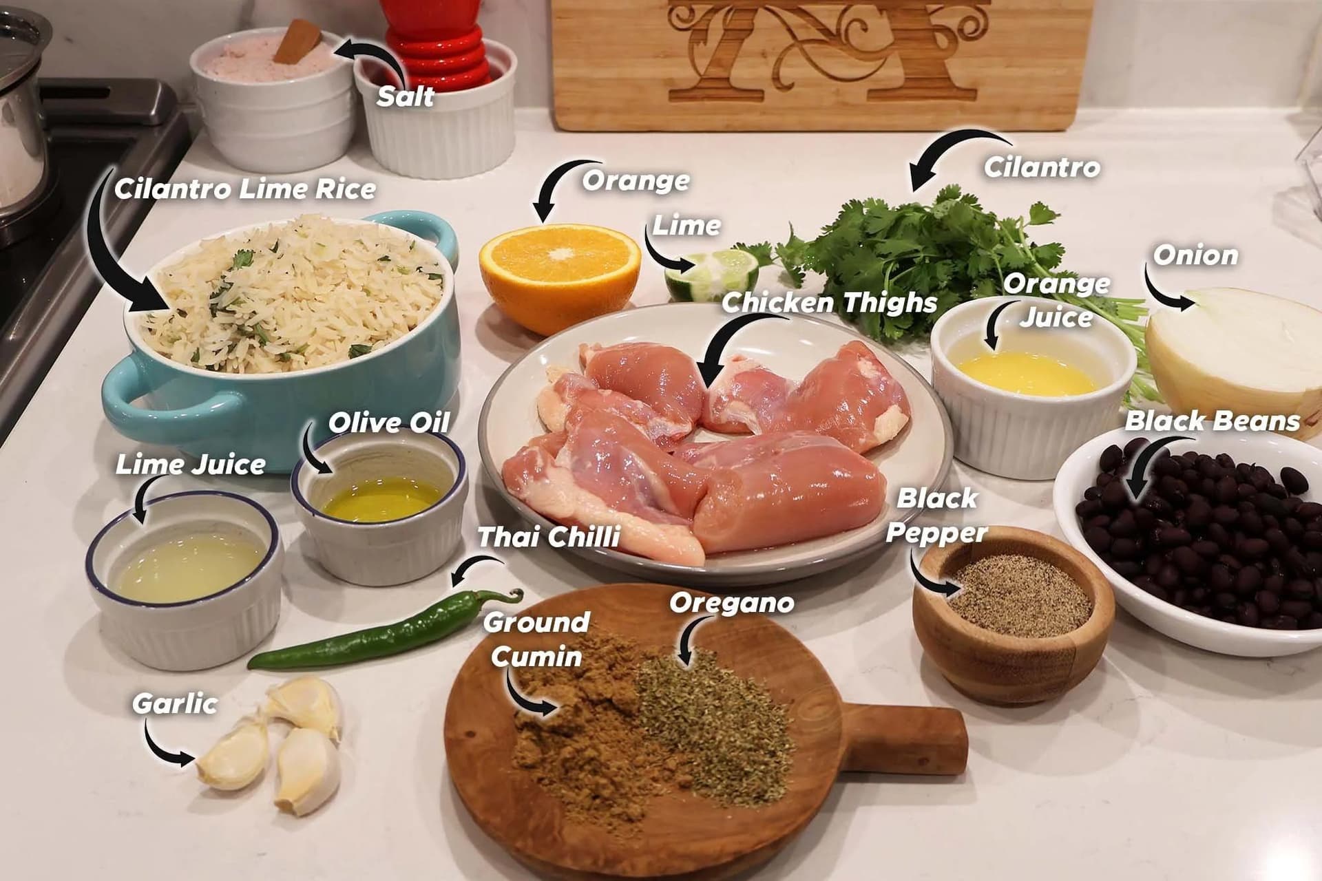 Ingredients for Cuban mojo chicken laid out on a white countertop, including chicken thighs, cilantro lime rice, citrus, spices, and beans.