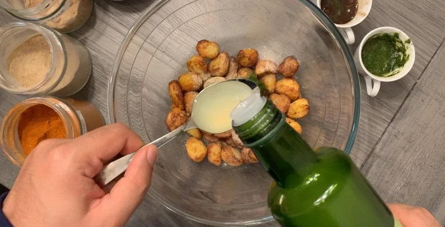 Hand pouring sauce over crispy golden air-fried baby potatoes in a glass bowl with spice jars nearby.