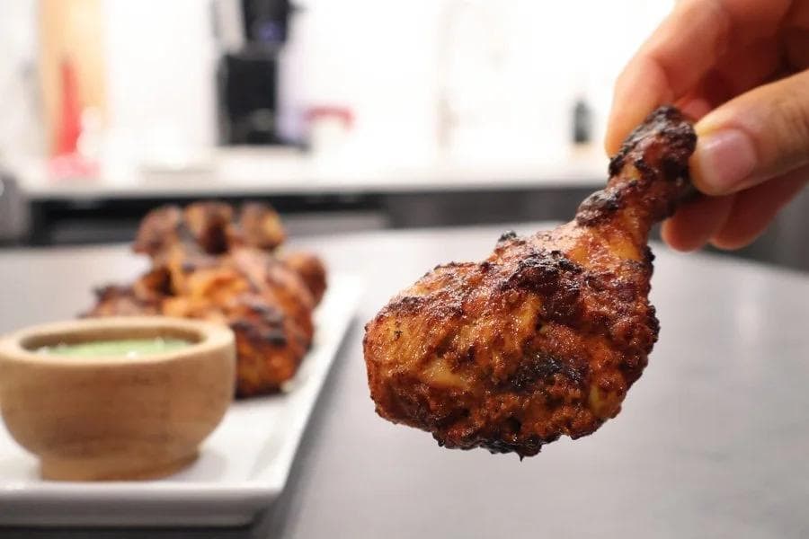 Hand holding a golden-brown tandoori chicken tikka piece with dipping sauce in background on white plate.