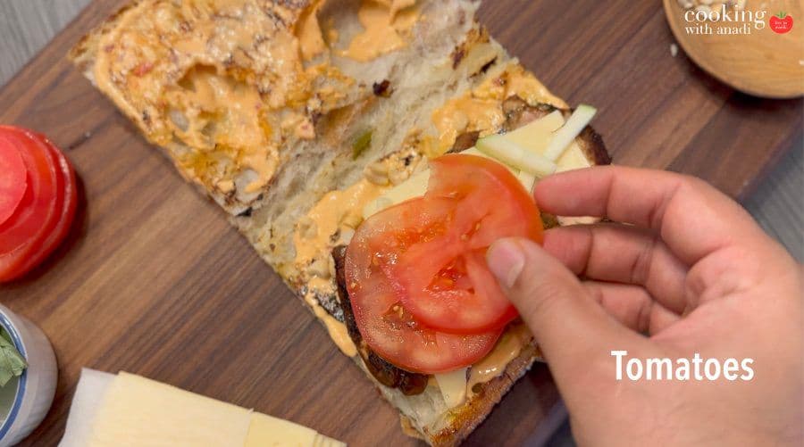 Hand placing tomato slices on Thai chicken sandwich with cheese and apple on toasted baguette.