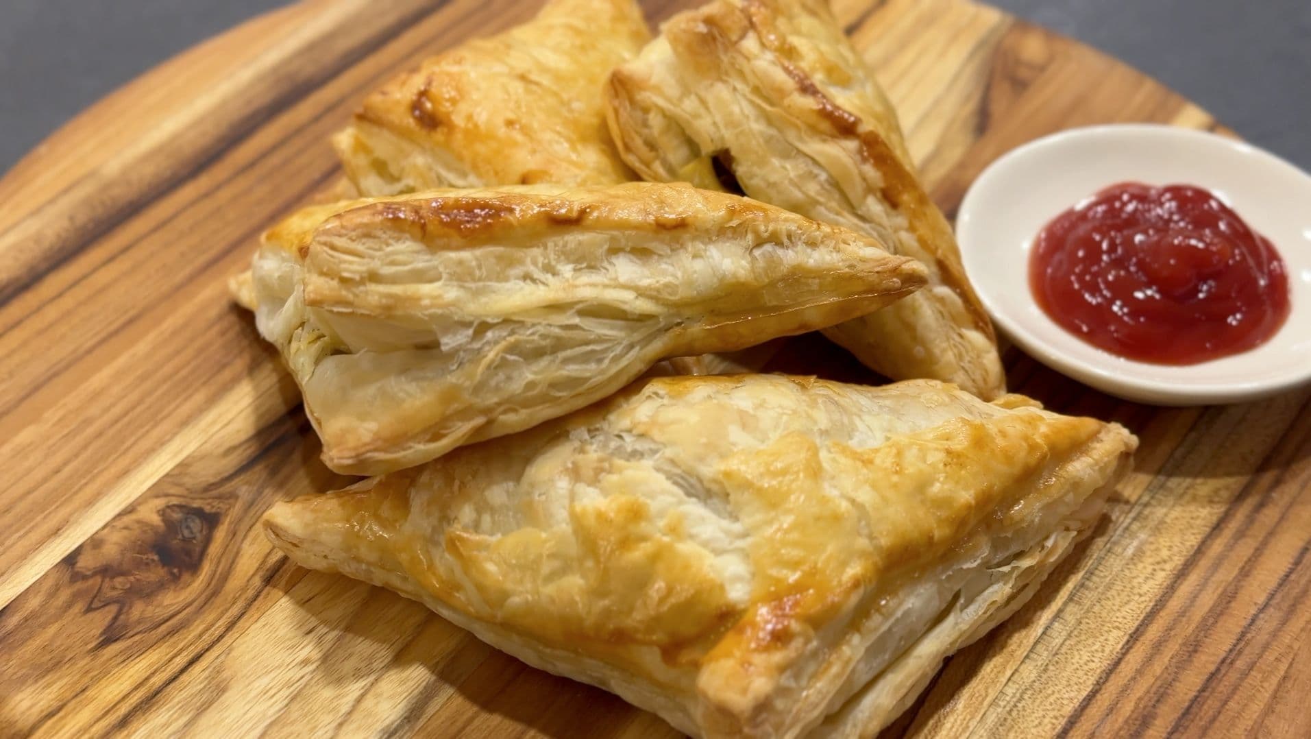Golden baked mushroom puff pastry patties on a wooden board with red jam sauce in a small bowl.