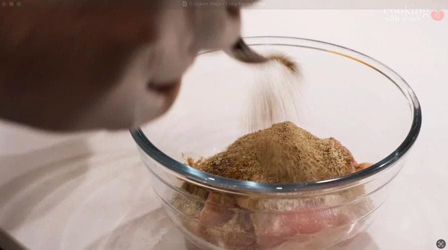 Cumin and other dry spices being sprinkled into a glass bowl with raw chicken pieces for marinade preparation.