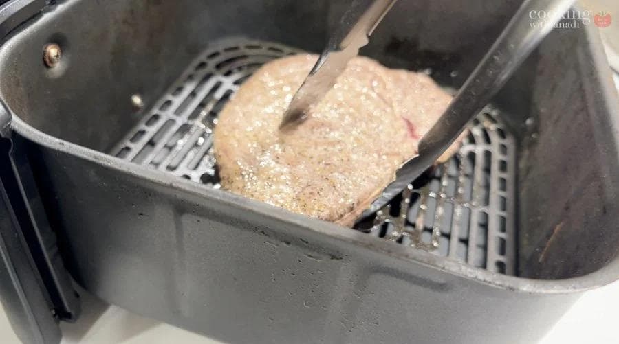 Raw sirloin steak being placed in air fryer basket with tongs for cooking.