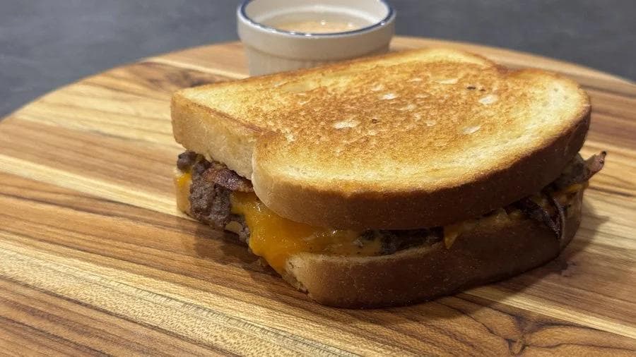 Smash burger patty melt on toasted bread with melted cheddar cheese and bacon, served on a wooden board with sauce.