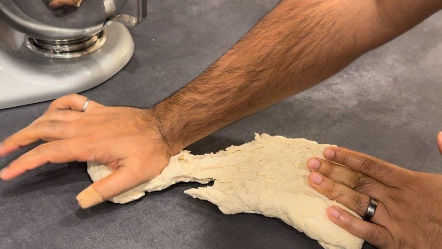 Hands kneading and stretching pizza dough on a dark countertop during manual dough preparation.