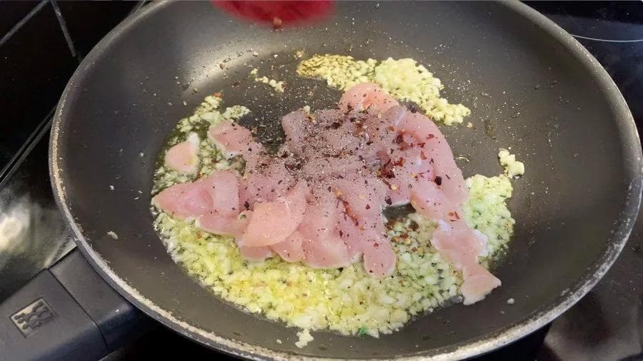 Diced chicken seasoned with chili flakes and black pepper cooking in a pan with sautéed garlic.