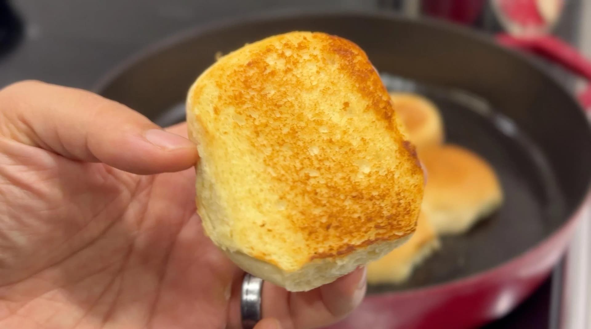 Toasted slider bun held in hand with golden-brown exterior, cast iron pan with additional buns visible in background