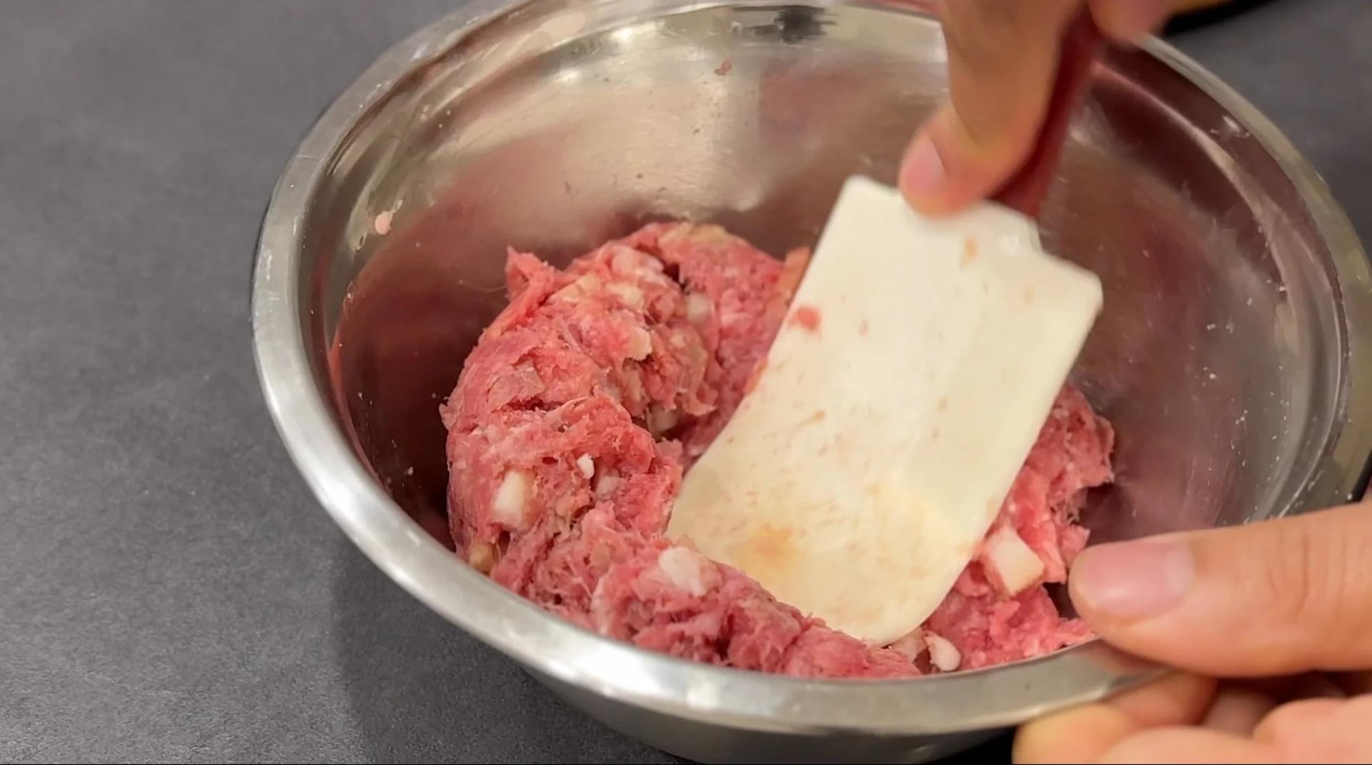 Ground beef mixture with bacon and seasonings in a bowl, butter being added by hand.