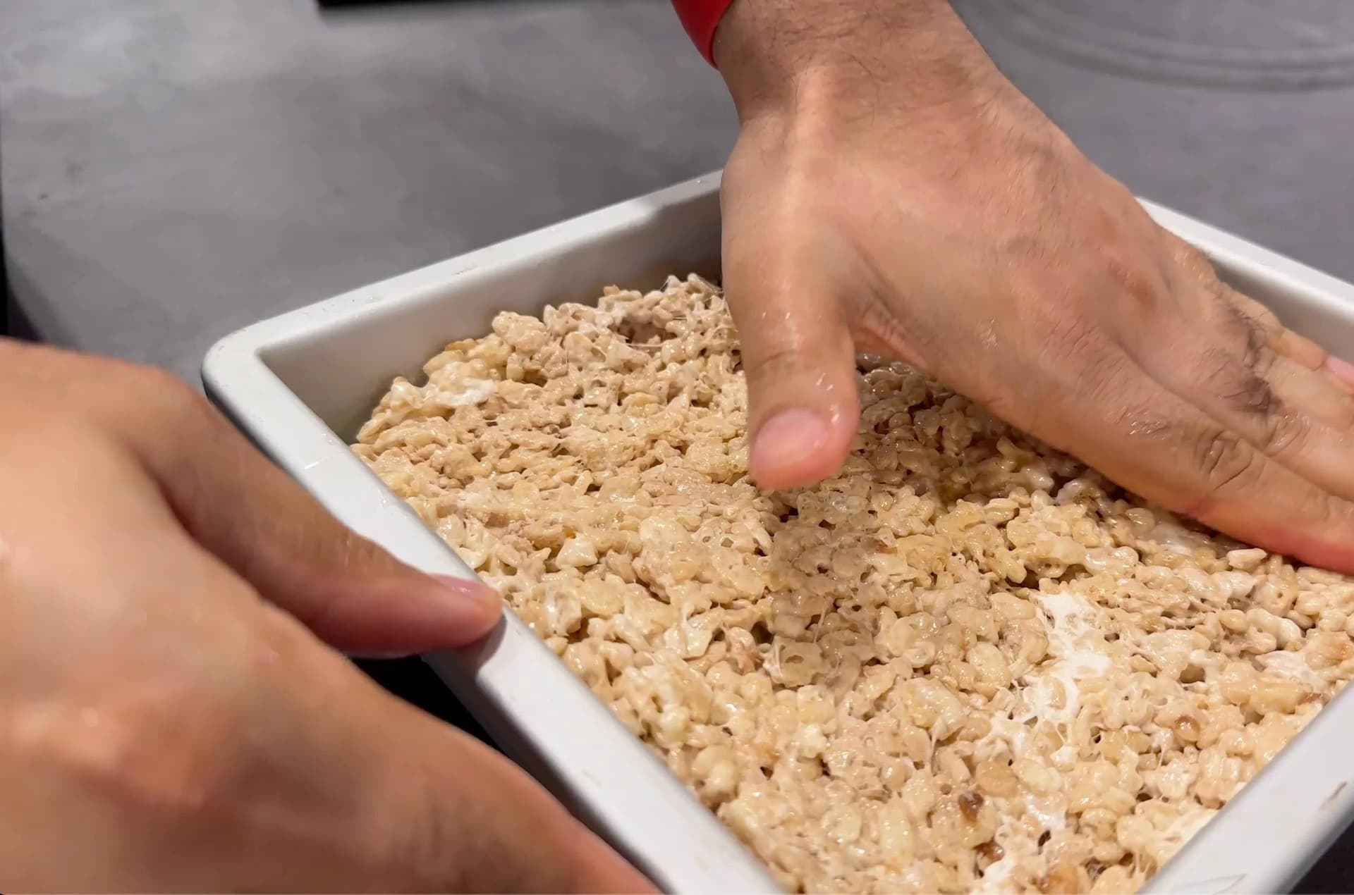 Hands patting down protein rice krispies mixture in white rectangular baking pan to flatten evenly.