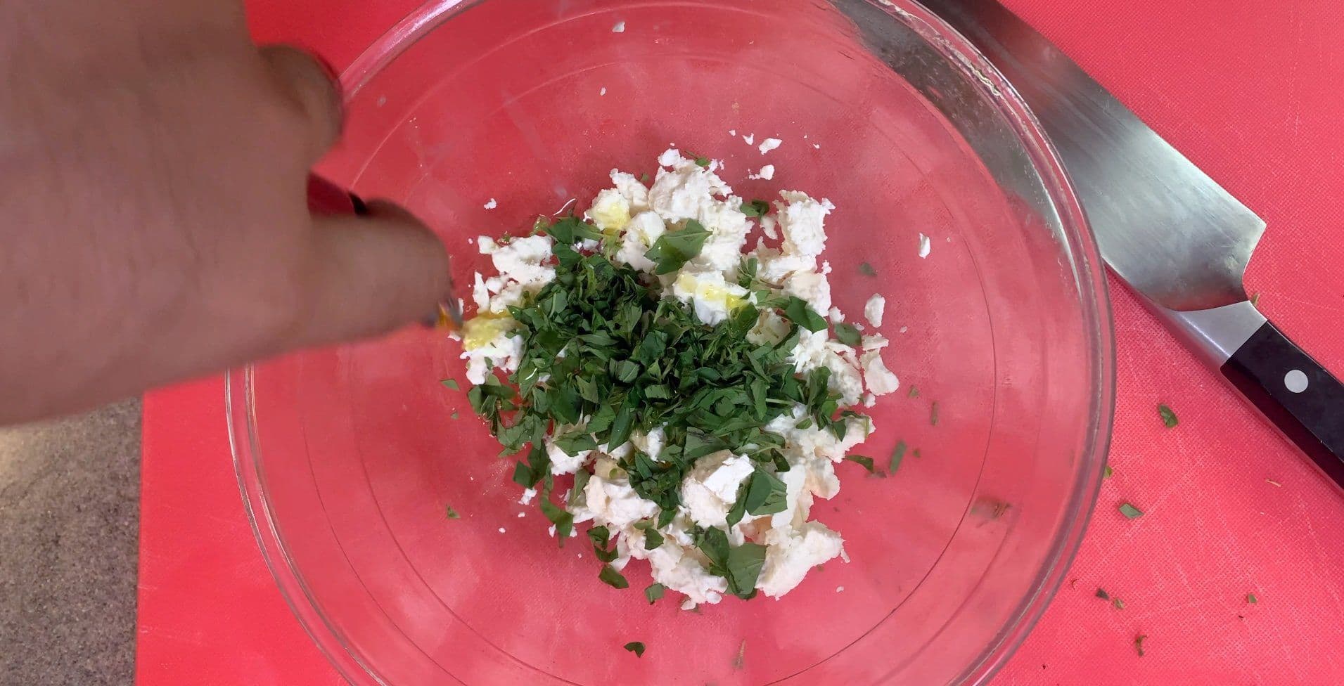 Crumbled feta cheese mixed with fresh oregano leaves, salt, and lemon zest in a clear mixing bowl.