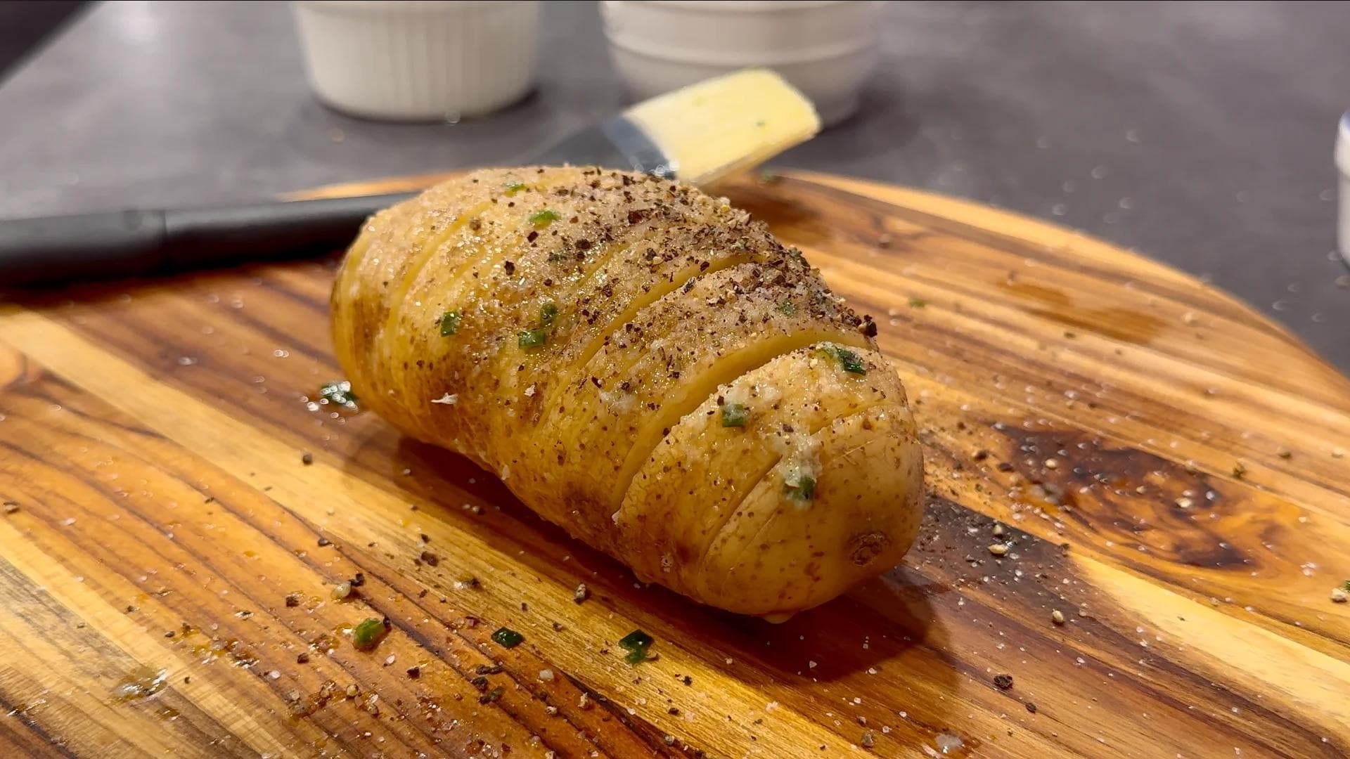 Cooked hasselback potato on wooden board, golden brown with herbs and black pepper seasoning, sliced partway through.