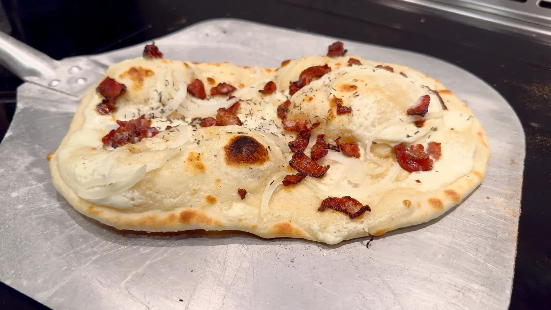 Broiled flammkuchen on parchment paper with crispy edges, melted cheese, and scattered bacon pieces.