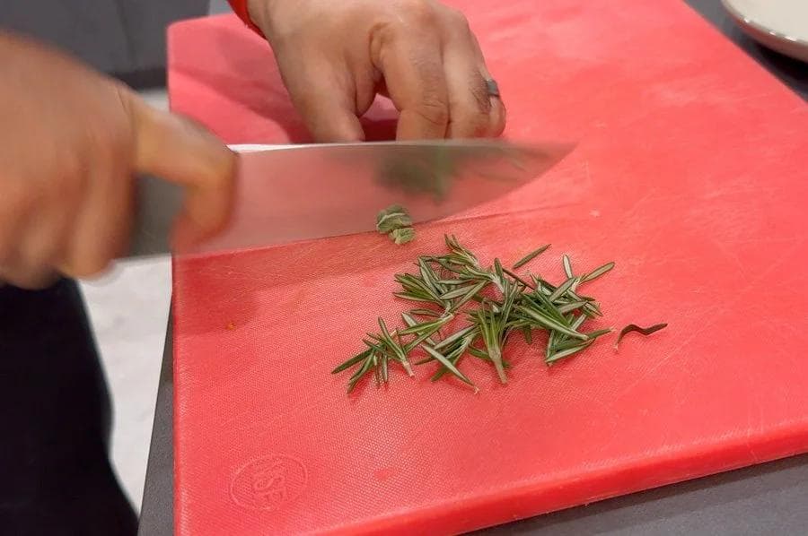 Chopping fresh rosemary herbs on a red cutting board with a chef's knife