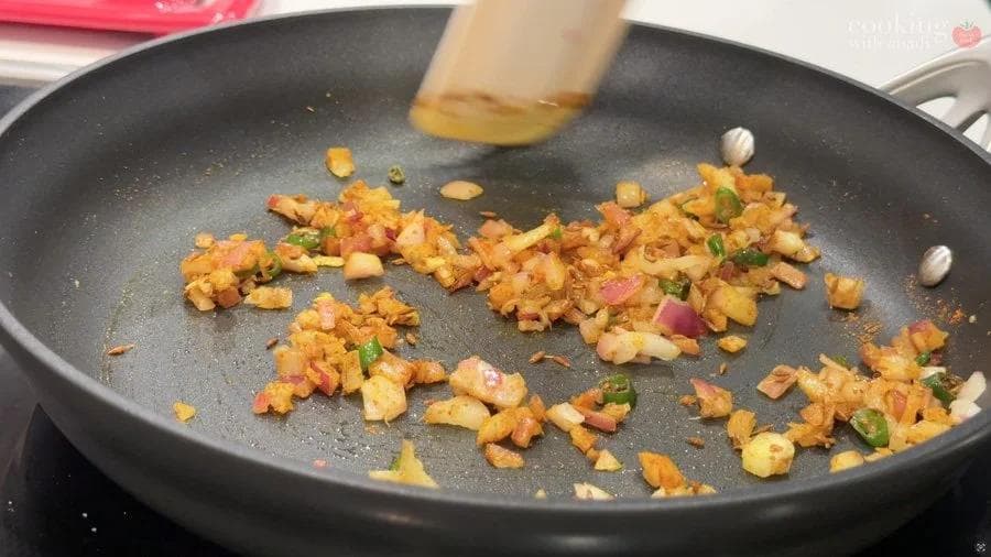 Diced onions and potatoes being cooked in a dark pan with spices and green chilies visible.