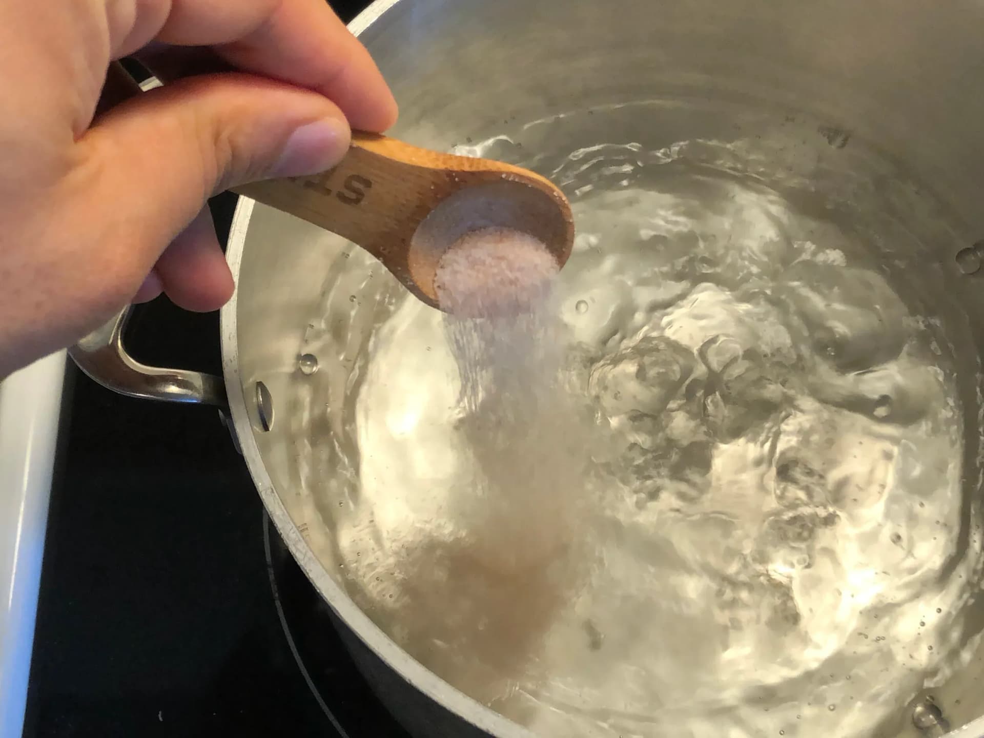 Hand pouring salt from wooden measuring spoon into boiling pasta water in pot.