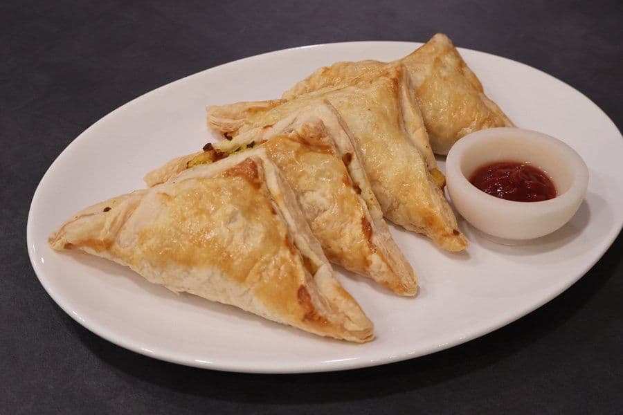 Three golden, crispy aloo puff pastry triangles on a white plate with a small bowl of red dipping sauce.
