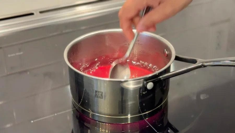 Pressing rose petals with a spatula in simmering red rose water in a saucepan on a stovetop.