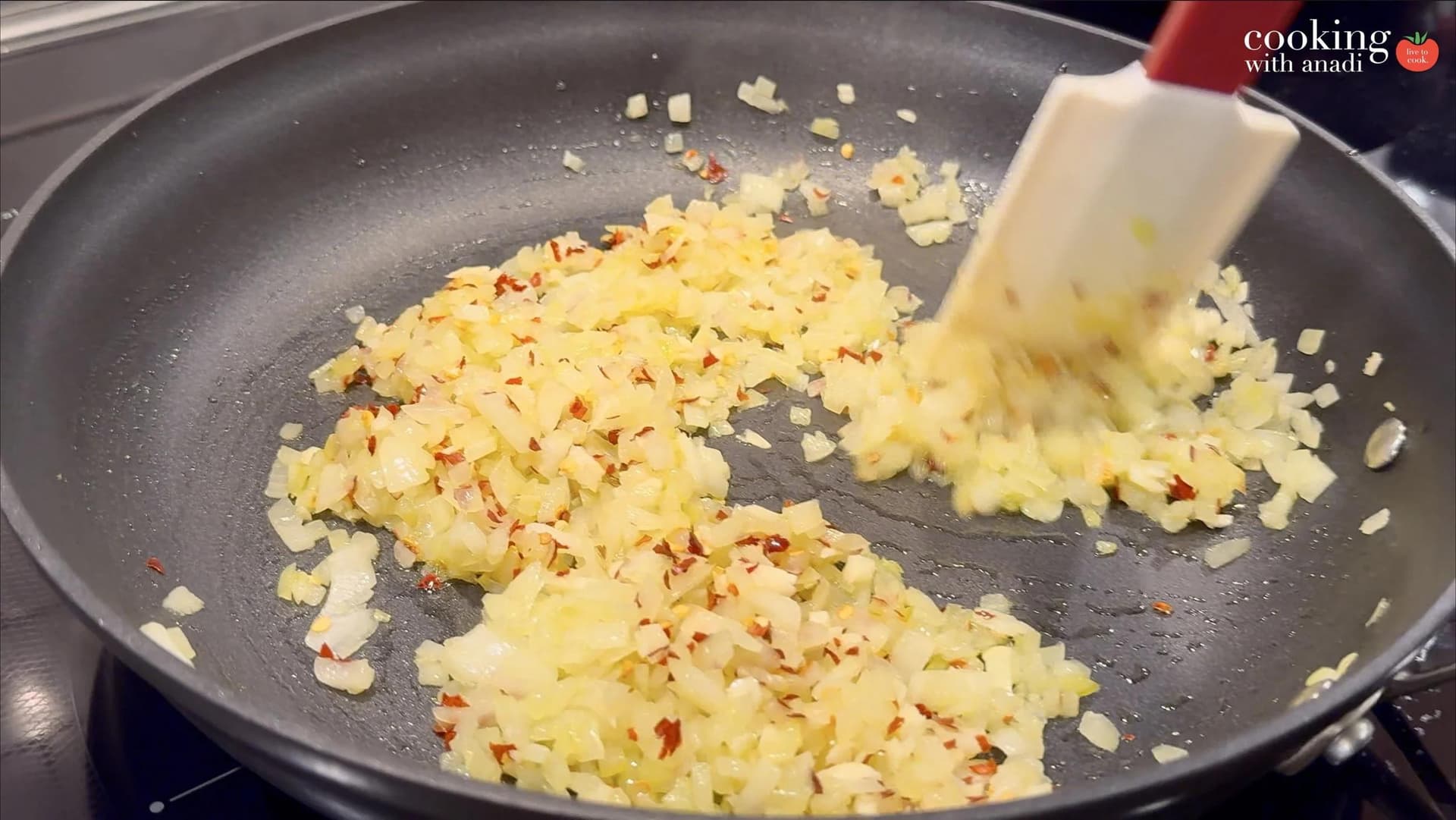 Diced onions and garlic with chili flakes sautéing in a nonstick pan, being stirred with a white silicone spatula.