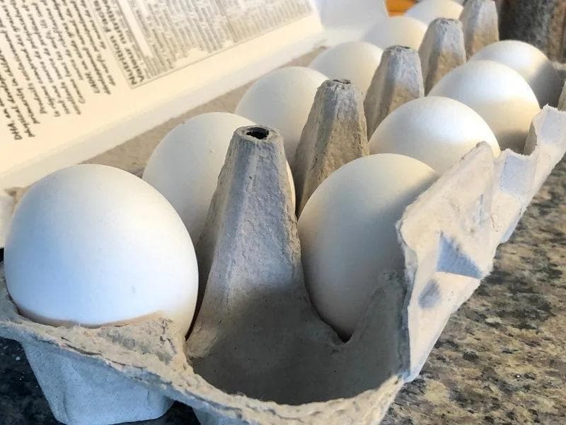 Raw eggs in a cardboard carton next to an open cookbook on a kitchen counter.