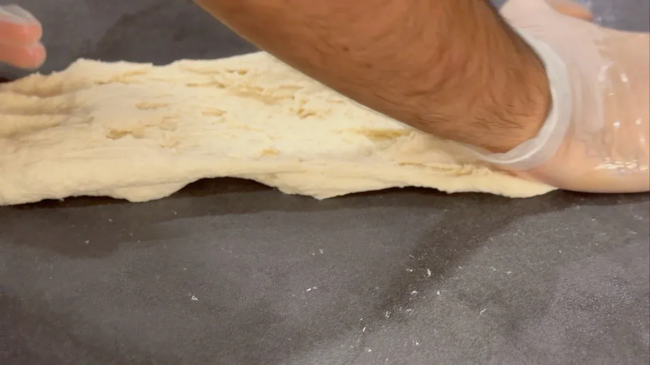 Hands kneading smooth, pale dough on a dark work surface dusted with flour.