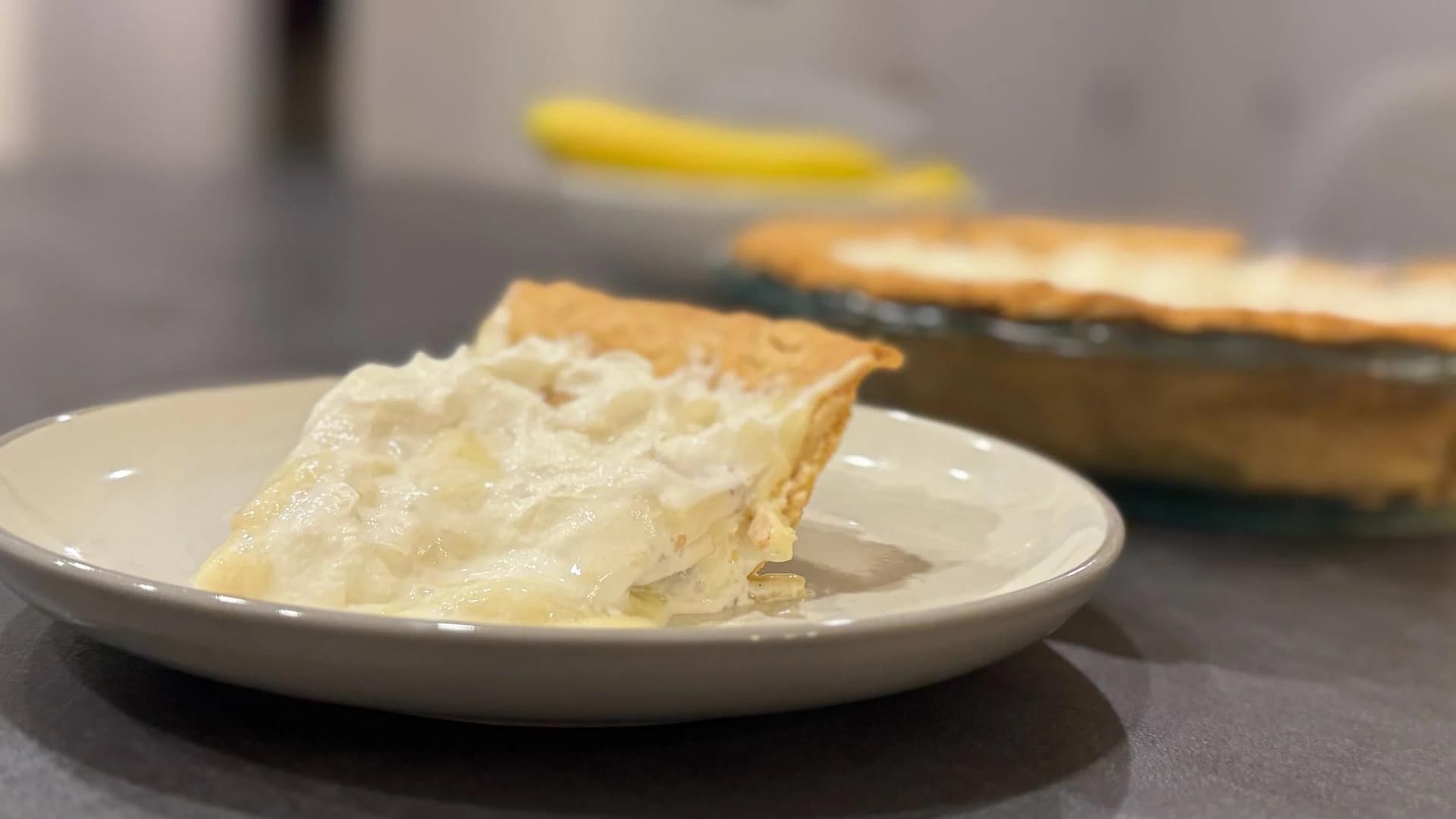Slice of no-bake banana cream pie with whipped cream topping on white plate, pie dish visible in background.